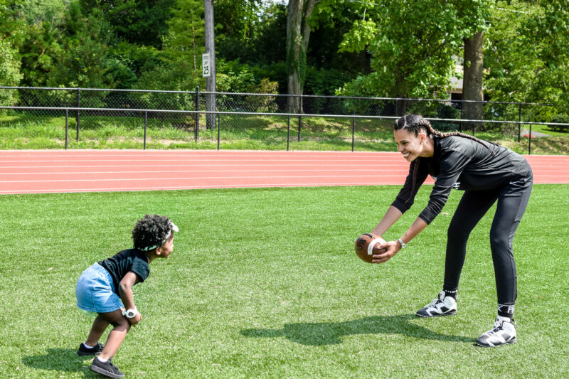 Mom and young daughter play football on a field together.