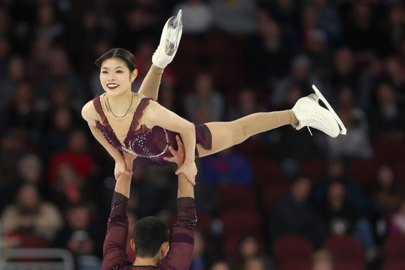 Female figure skater is hoisted in the air by her partner on the ice as she smiles out at the crowd.