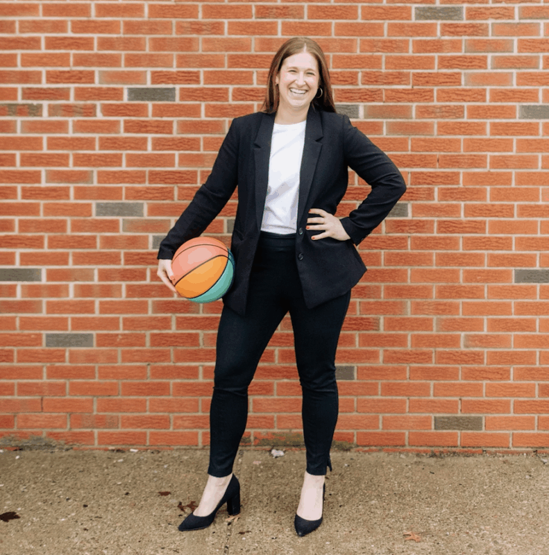 A woman in a black suit holding a basketball standing in front of a brick background.