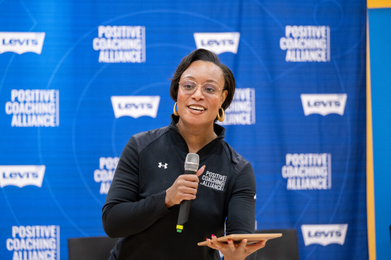 A woman wearing a black track jacket holding a microphone and an ipad speaking at an event.