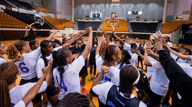 A group of young girls gather in a huddle on a basketball court with their hands in the air.
